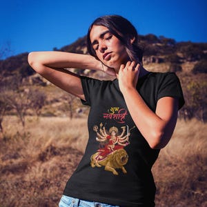 May include: Black t-shirt with a vibrant graphic of a deity on a lion, and text in a foreign script. The design uses red, gold, and orange hues. The shirt is worn with blue jeans.