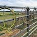 May include: A metal fence with several coiled ropes hanging from it. The ropes are various colors, including yellow, brown, and red. The background features a green field and a cloudy sky.