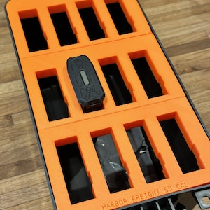 Puede incluir: Inserto de espuma naranja con múltiples compartimentos para guardar objetos pequeños. El inserto está en una caja de almacenamiento de plástico negro con el texto "HARBOR FREIGHT 50 CAL" impreso en el lateral.