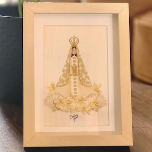 May include: Framed artwork depicting a religious figure in a gold crown and robe, surrounded by white flowers. The artwork is in a light wood frame, resting on a wooden surface.