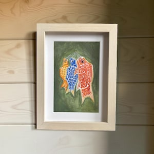 May include: Framed artwork featuring three colorful carp-shaped windsocks, a traditional Japanese symbol. The carp are orange, blue, and red, set against a green background. The artwork is in a light-colored wooden frame, suitable for home decor.