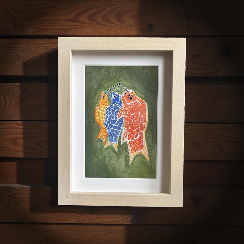 May include: A framed watercolor painting of three colorful carp kites, in orange, blue, and red, hanging against a green background. The artwork is displayed in a light wood frame, perfect for home decor.