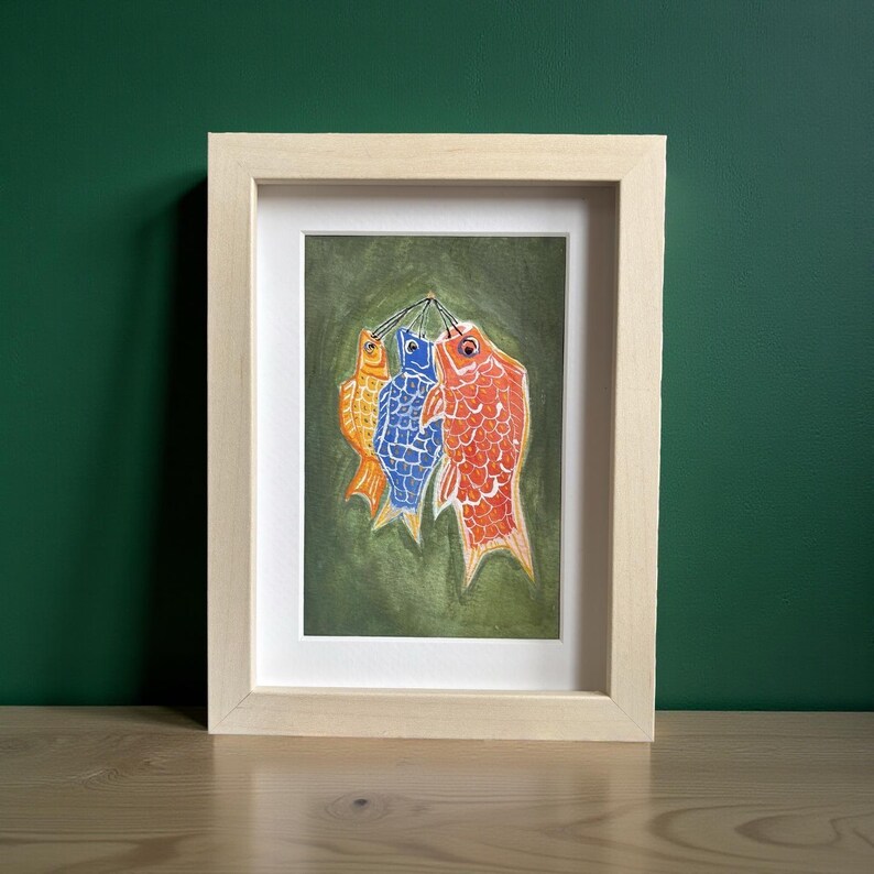 May include: Framed artwork featuring three colorful fish hanging against a green background. The fish are orange, blue, and red, with detailed scale patterns. The artwork is in a light-colored wooden frame, suitable for wall decor.