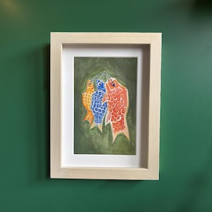 May include: A framed artwork featuring three colorful fish-shaped windsocks. The fish are orange, blue, and red, with detailed scale patterns. The artwork is set against a green background and framed in a light wood frame.
