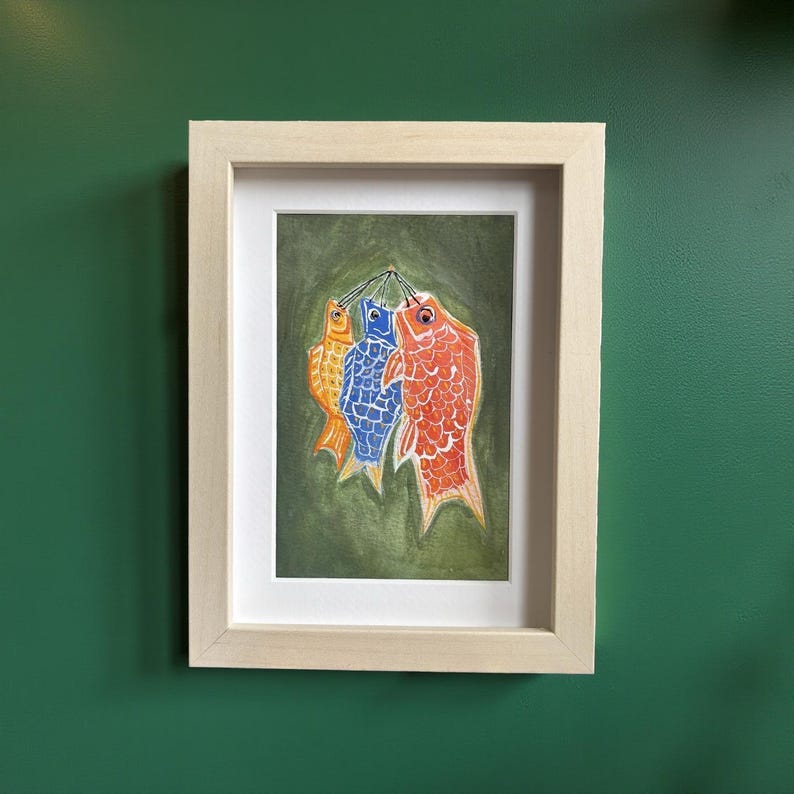 May include: A framed artwork featuring three colorful fish-shaped windsocks. The fish are orange, blue, and red, with detailed scale patterns. The artwork is set against a green background and framed in a light wood frame.