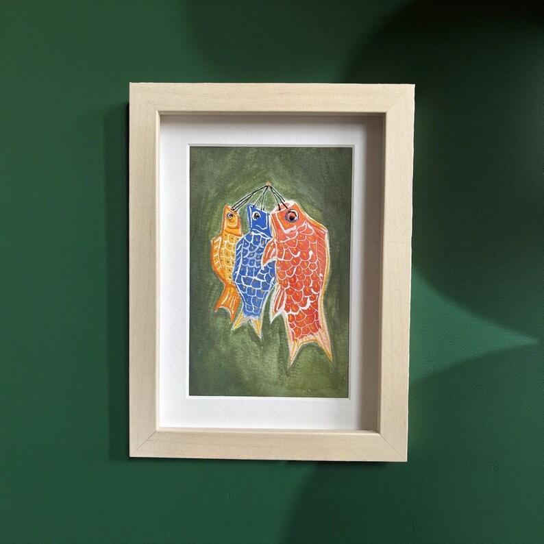May include: A framed artwork featuring three colorful fish hanging against a green background. The fish are orange, blue, and red, with detailed scale patterns. The artwork is in a light wood frame, displayed on a dark green wall.