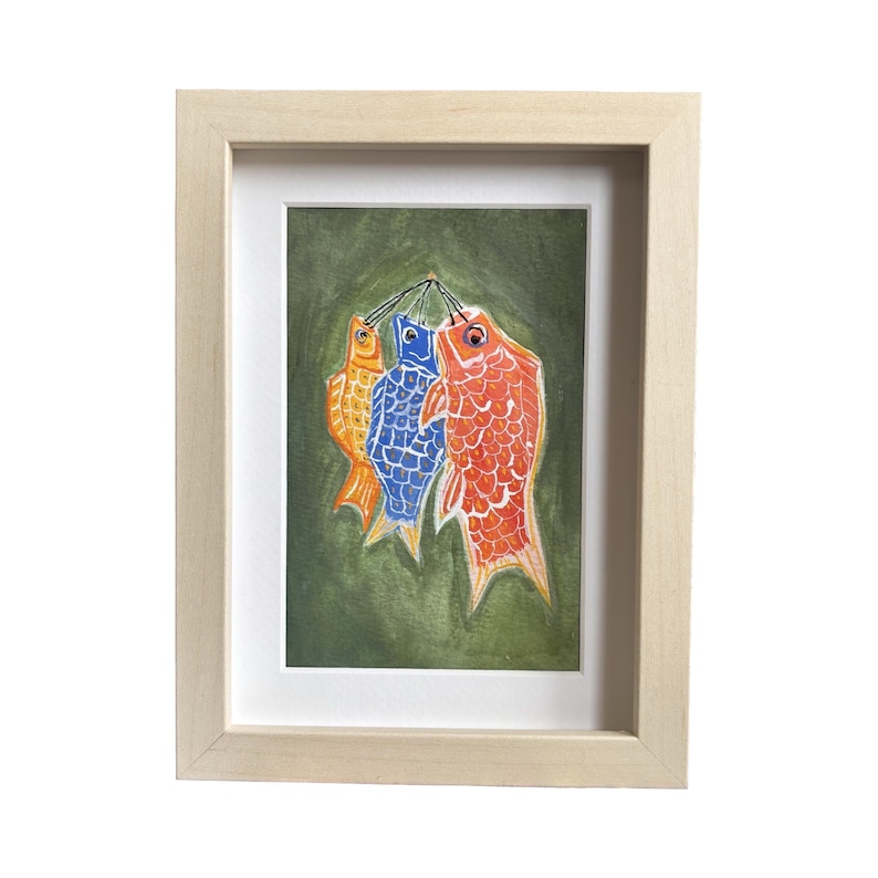 May include: Framed artwork featuring three colorful carp streamers, a traditional Japanese symbol. The carp are orange, blue, and red, hanging against a green background. The artwork is in a light wood frame with a white mat.