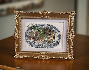 May include: A decorative framed artwork with a gold-coloured ornate frame. The artwork inside features a blue and white patterned border, and a central display of dried leaves, pine needles, and a rose.