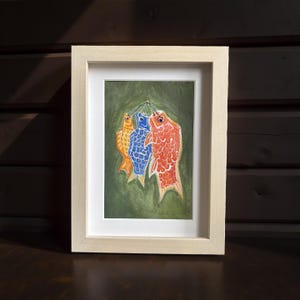 May include: Framed artwork featuring three colorful fish hanging against a green background. The fish are orange, blue, and red, with detailed scale patterns. The artwork is in a light-colored wooden frame, suitable for home decor.