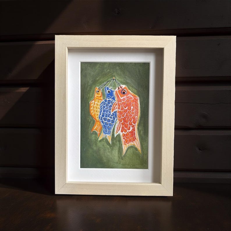 May include: Framed artwork featuring three colorful fish hanging against a green background. The fish are orange, blue, and red, with detailed scale patterns. The artwork is in a light-colored wooden frame, suitable for home decor.