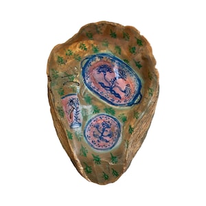 May include: A brown seashell with a painted design of blue and pink plates and a glass. The shell is decorated with green leaves.