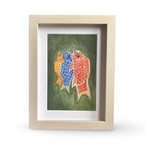 May include: A framed artwork featuring three colorful fish-shaped windsocks. The fish are orange, blue, and red, with detailed scale patterns. The background is a textured green, and the frame is light wood.