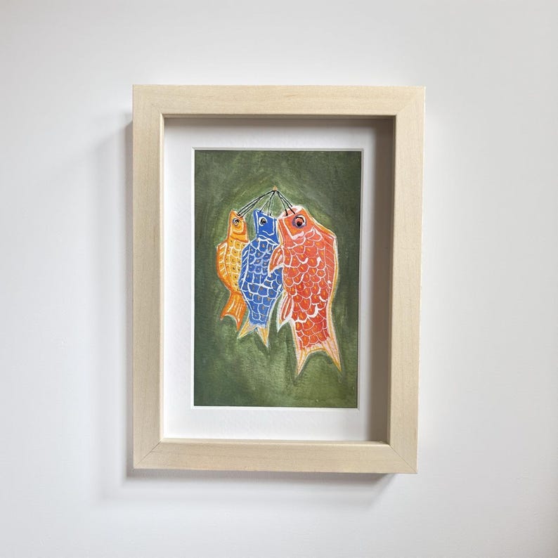 May include: Framed artwork featuring three colorful fish hanging against a green background. The fish are orange, blue, and red, with detailed scale patterns. The frame is a light wood color, adding a natural touch to the art.