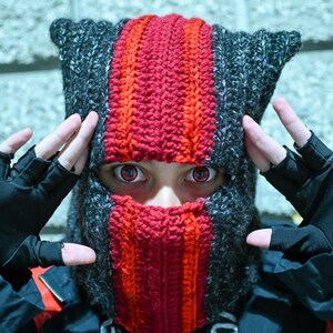 Könnte beinhalten: Handgestrickte Sturmhaube in Grau und Rot. Die Sturmhaube hat einen zentralen roten Streifen und eine passende rote Gesichtsbedeckung. Die Person trägt schwarze fingerlose Handschuhe und rote Kontaktlinsen.