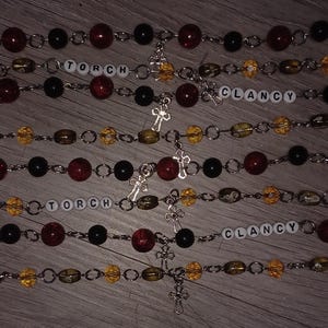 Op de afbeelding: Verschillende armbanden met rode, zwarte en gele kralen, en zilverkleurige schakels. Sommige armbanden hebben de namen "TORCH" en "CLANCY" in witte letterkralen. Elke armband heeft een kleine kruisbedel.
