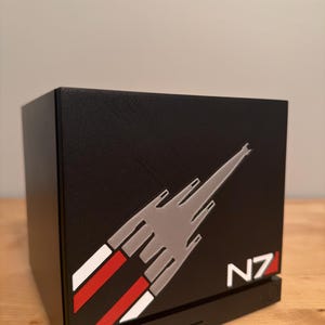 May include: A black cubic computer case featuring a gray, white, and red graphic of a spaceship silhouette on the front panel. The text N7 is printed in white and red in the bottom right corner. The base includes two USB ports and a power button.