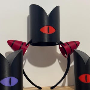 May include: Black crown-shaped headbands with red and purple eye designs. The headbands have a black base with a red eye design. The side pieces are red with black stripes. The eye designs are red and purple.