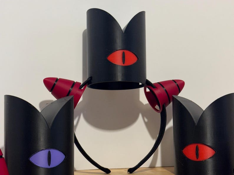 May include: Black crown-shaped headbands with red and purple eye designs. The headbands have a black base with a red eye design. The side pieces are red with black stripes. The eye designs are red and purple.