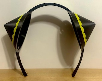 May include: Black headband with two cone-shaped ear accessories. Each ear is black with a yellow scalloped trim. The headband and ear attachments are made of a flexible material. The design is simple and modern.