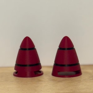 May include: Two maroon-colored, cone-shaped objects with black bands. The objects have a rounded top and a base with cut-out sections. They are placed on a light brown wooden surface.