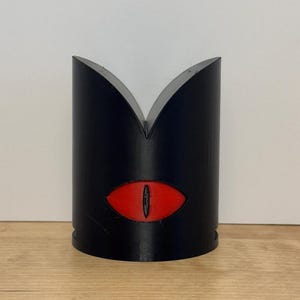 May include: A black, cylindrical object with a red eye design. The top of the object has a V-shaped cutout. The eye is oval-shaped with a black pupil. The object is likely a decorative item or a container.