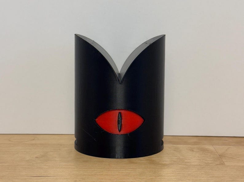 May include: A black, cylindrical object with a red eye design. The top of the object has a V-shaped cutout. The eye is oval-shaped with a black pupil. The object is likely a decorative item or a container.