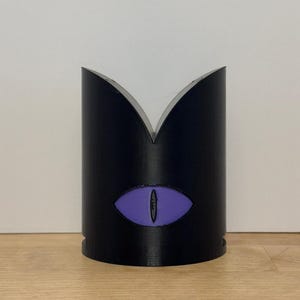 May include: A black cylindrical object with a purple eye design. The top of the cylinder has a V-shaped cutout. The eye is centered on the front of the cylinder. The object is sitting on a wooden surface.