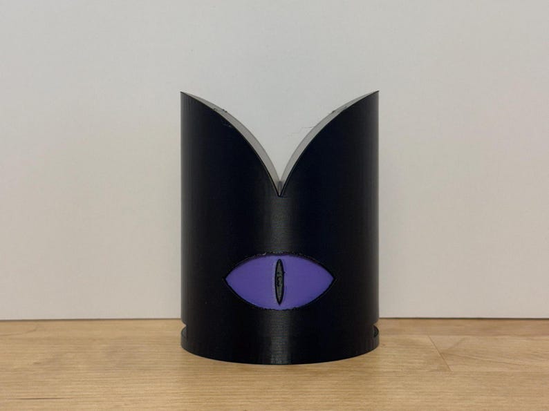 May include: A black cylindrical object with a purple eye design. The top of the cylinder has a V-shaped cutout. The eye is centered on the front of the cylinder. The object is sitting on a wooden surface.