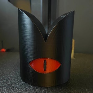 May include: A black cylindrical container with a textured surface and a stylized red eye design. The top of the container has a pointed, crown-like shape. The eye is oval with a black pupil.