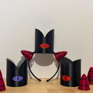 May include: Black headbands with red and black horns and black crowns with eye designs in red and purple. Several red and black cone-shaped objects are also present. The items are arranged on a wooden surface.