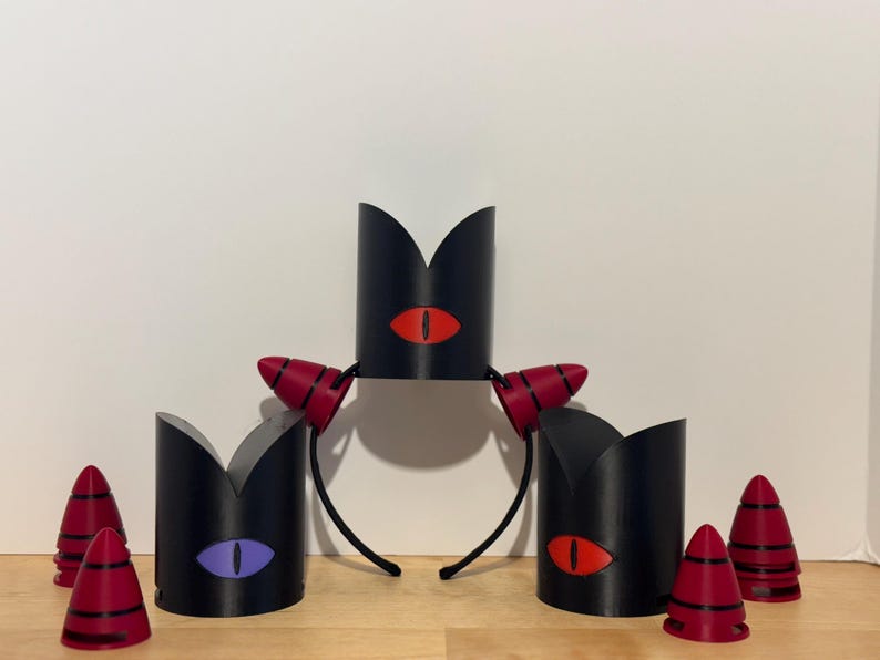 May include: Black headbands with red and black horns and black crowns with eye designs in red and purple. Several red and black cone-shaped objects are also present. The items are arranged on a wooden surface.