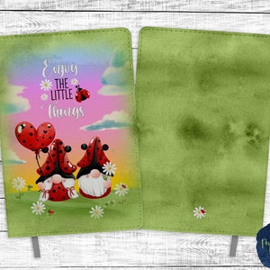 May include: A green journal cover with a watercolor background. The front cover features two gnomes with red and black hats, a red heart balloon, and the text "Enjoy the little things".