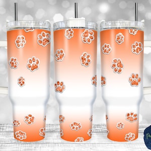 Op de afbeelding: Drie roestvrijstalen bekers met een witte achtergrond en een oranje pootprintontwerp. De pootprints hebben een glittereffect.