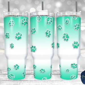 May include: Three stainless steel tumblers with a teal and white ombre design and glitter paw prints. The tumblers have a handle and a lid.