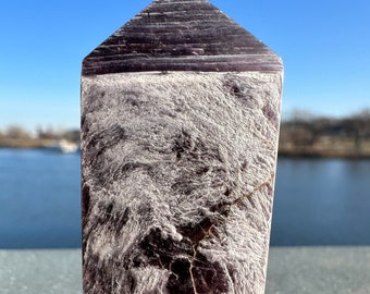 May include: A purple and white crystal point with a striated pattern. The crystal is sitting on a grey surface.