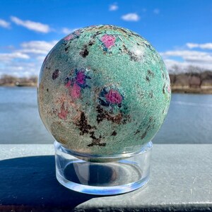 May include: A green and pink sphere with black and brown inclusions, resting on a clear acrylic stand. The sphere is a polished gemstone, likely a variety of jasper.