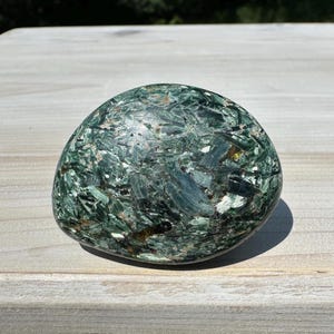 May include: A rounded, polished stone with a mottled appearance. The stone's surface is primarily a deep, forest green, interspersed with lighter green and white crystalline inclusions. The stone is smooth and appears to be a natural mineral specimen.