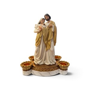 May include: A decorative figurine depicting a family of three figures in cream and gold robes standing on a circular base. The base features a white marble-like finish with gold trim and four integrated candle holders with scalloped edges.