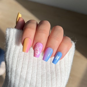 K&ouml;nnte beinhalten: Nahaufnahme einer Hand mit buntem Ombre-Nagellack. Die N&auml;gel sind mit wei&szlig;en Schmetterlings- und Herzmotiven verziert.