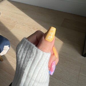 K&ouml;nnte beinhalten: Nahaufnahme einer Hand mit langen, gelben N&auml;geln. Die N&auml;gel sind mit einem wei&szlig;en Design verziert, das "HO" und ein geometrisches Muster zeigt. Die Hand tr&auml;gt einen wei&szlig;en Pullover.