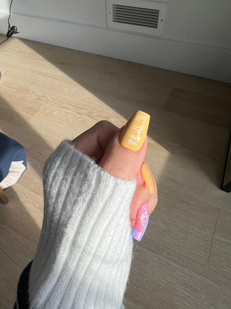 K&ouml;nnte beinhalten: Nahaufnahme einer Hand mit langen, gelben N&auml;geln. Die N&auml;gel sind mit einem wei&szlig;en Design verziert, das "HO" und ein geometrisches Muster zeigt. Die Hand tr&auml;gt einen wei&szlig;en Pullover.
