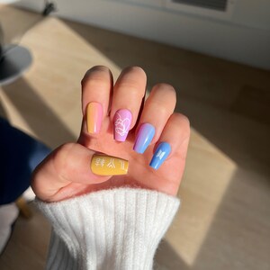 K&ouml;nnte beinhalten: Nahaufnahme einer Hand mit langen, bunten N&auml;geln. Die N&auml;gel sind in Gelb-, Rosa- und Blaut&ouml;nen lackiert und mit wei&szlig;en Designs verziert. Das Design auf dem gelben Nagel lautet "방탄소년단" in Koreanisch.