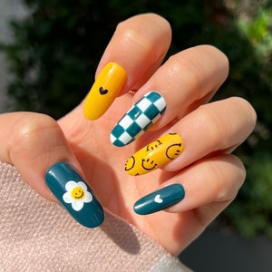 Könnte beinhalten: Nahaufnahme einer Hand mit blaugrünem und gelbem Nagellack. Die Nägel sind mit einem karierten Muster, Smiley-Gesichtern und einer Gänseblümchen verziert.