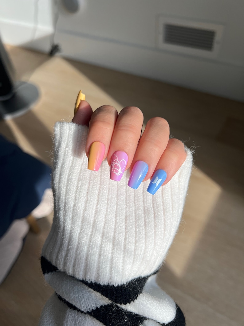 K&ouml;nnte beinhalten: Nahaufnahme einer Hand mit buntem Ombre-Nagellack. Die N&auml;gel sind mit wei&szlig;en Schmetterlings- und Herzmotiven verziert.