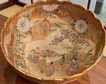 Könnte beinhalten: Eine große, runde Keramikschale mit gewelltem Rand, verziert mit handgemalten Szenen von Figuren in traditioneller Kleidung in einer Landschaft, umgeben von detaillierten Blumenmotiven, Bambusstangen und goldfarbenen Zierrändern.