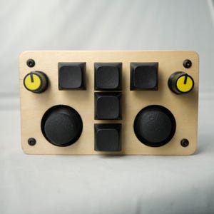 May include: A wooden control panel with black buttons, knobs with yellow detailing, and black joysticks. The panel has a light wood finish and is secured with black screws. The design is symmetrical, with controls arranged in a grid.
