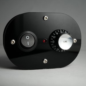 Puede incluir: Un panel de control negro con un interruptor de palanca, un mando de tono y una luz indicadora roja. El mando de tono tiene una tapa plateada y está etiquetado como "TONE". El panel está asegurado con cuatro tornillos plateados.