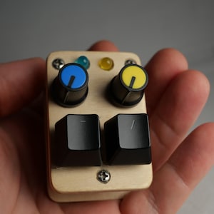 Puede incluir: Un pequeño dispositivo rectangular de madera con dos perillas negras con marcas azules y amarillas, y dos botones cuadrados negros. El dispositivo también tiene dos pequeñas luces de colores y está asegurado con tornillos.