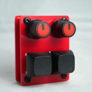 Puede incluir: Un dispositivo electrónico rojo con dos perillas negras con indicadores rojos y dos botones cuadrados negros. El dispositivo está montado con tornillos negros.