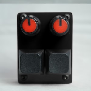 Puede incluir: Un dispositivo electrónico negro con dos perillas rojas y negras y dos botones cuadrados negros. Las perillas tienen un centro rojo con un indicador blanco. El dispositivo está montado en una placa negra con tornillos.